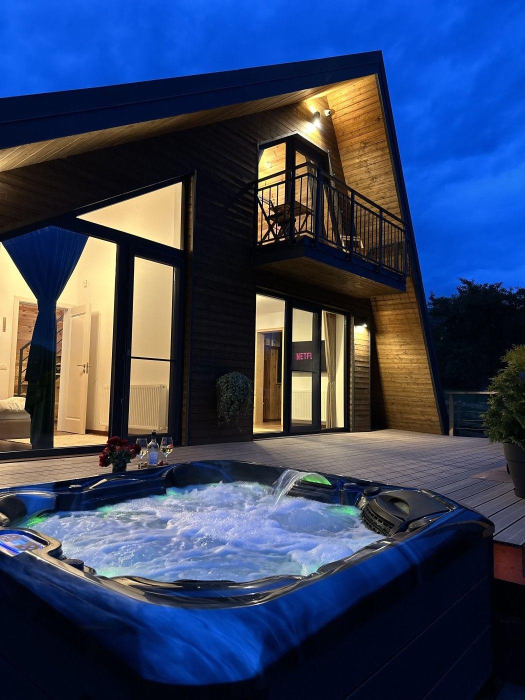 Modern wooden house with large windows, balcony, and outdoor hot tub on a deck at night.