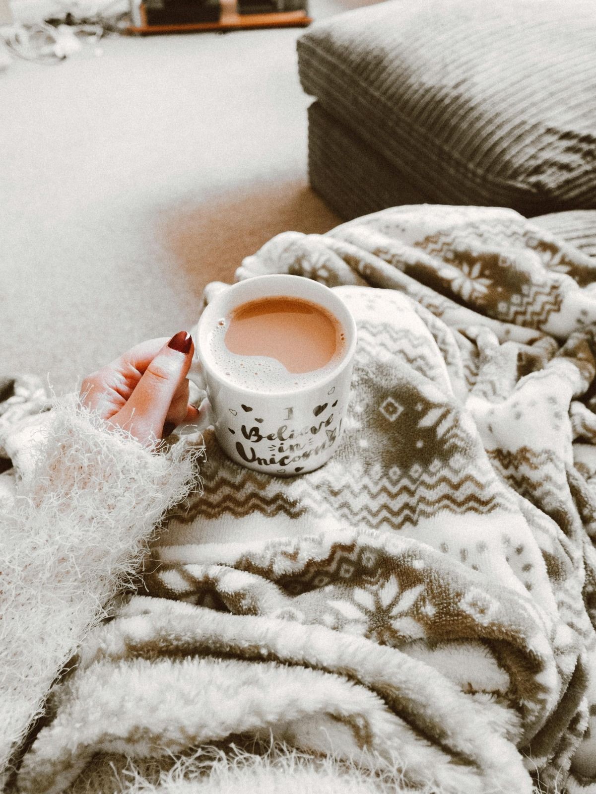 Person holding a mug of tea while wrapped in a patterned blanket, sitting on a fluffy couch.