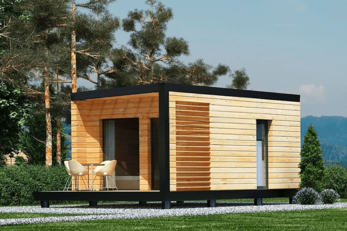 Modern small wooden house with large windows and outdoor seating, set in a forested area.