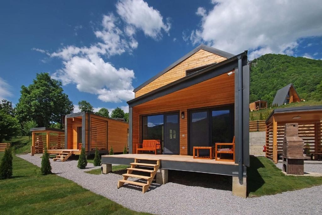 Modern wooden cabin with large windows and porch, surrounded by greenery and a mountain in the background.