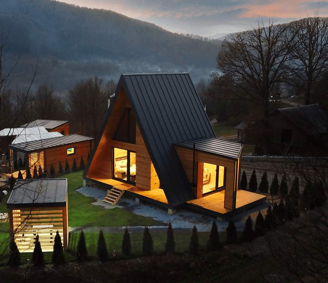 A-frame wooden cabin with large windows, illuminated at dusk, surrounded by trees and hills.