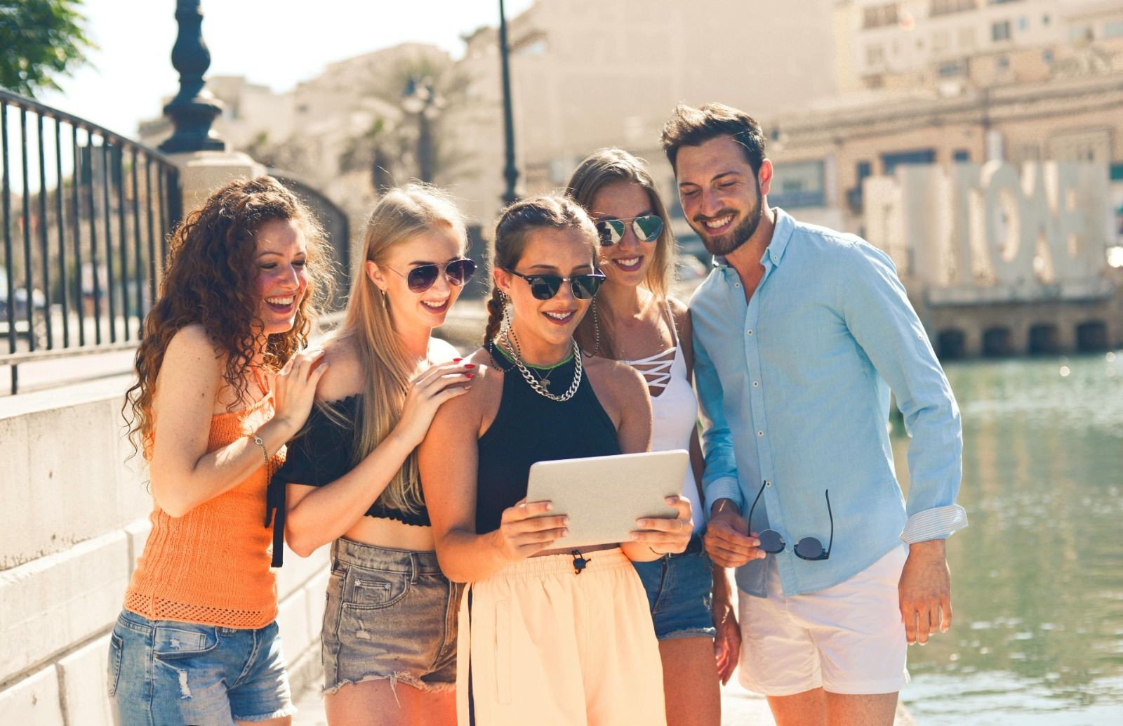 Group of friends outdoors, smiling and looking at a tablet by the water.